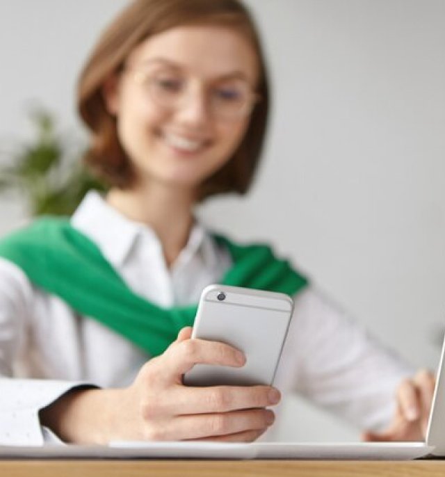 Lächelnde Frau nutzt ihr Smartphone und ihren Laptop, symbolisiert einfache Online-Konsultationen und nahtlose Koordination von Medizintourismus mit ROYAL MEDICAL.