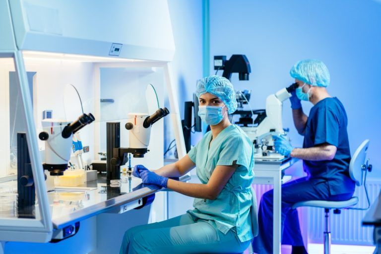 Two IVF scientists working with microscopes in a modern laboratory, wearing surgical scrubs, masks, and hair covers.
